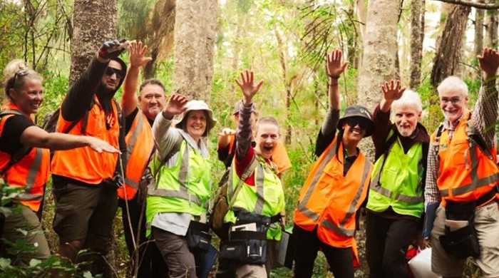 Eight people in high vis gear stand together in the forest holding one arm up, hand outstretched.