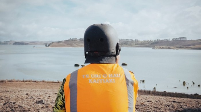 A person sits with their back to the camera, wearing a high vis vest and helmet, looking out over a body of water.